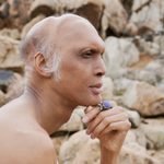 A person with medium skin rests their chin on their hand in front of a tan, rocky landscape.