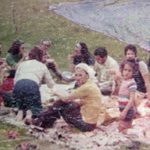 An old and restored image of adults and children sitting and eating at a picnic blanket on grass at a park.
