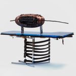 A blue acrylic tabletop resting on a metal chair with leather straps. A brown ovoid shape with a large stick behind it sits atop the table.