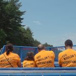 Six people in a blue boat wear the same yellow tshirt that reads “YOU WOULDN’T UNDERSTAND...” on the back. The water is surrounded by trees.