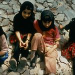 An image looking down toward four kids with dark hair, medium skin tone, and headscarves, sitting on rocks and holding apples.