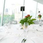 Round dining tables with white table cloth bearing silverware, wine glasses, plant center piece, and a black table sign reading "6" in front of a large window during the daytime.