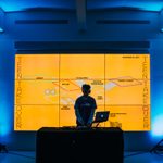 A DJ stands at a table with his gear in front of an orange screen reading "Teen Takeover" in a blue light room with speakers on stands