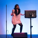 A singer dances on stage with a microphone and a monitor in front of a blue background