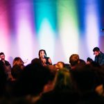 A band with a lead singer performs to an audience in front of a multicolored light stage