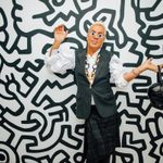 An adult poses in front of a black and whtie Keith Herring inspired wall, holding a leather skull bag, wearing daisy glasses and a fabulous outfit