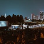 Crowds of people on pinic blankets on a hillside next to a museum watch a film projected on a large outdoor screen with a band playing in front of it and the Minneapolis skyline in the background.