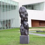 A large bronze sculpture that looks like a figure, stands upright in front of a white building outside on the lawn.