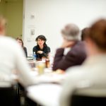 A group of adults sit and listen in a conference room at a table filled with papers and beverages