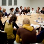 A group of adults sit and listen in a conference room at a table filled with papers and beverages