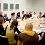 A group of adults sit and listen in a conference room at a table filled with papers and beverages