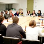 A group of adults sit and listen in a conference room at a table filled with papers and beverages