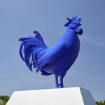 A large blue rooster sculpture sits on top of a concrete slab outdoors, blue sky in the background.