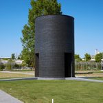 A large black brick circular building with doors on each side sits in a grassy lawn with concrete pathways leading to the doors.