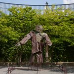 A large metal sculpture, loosely human figure shaped stands in front of a wall of trees outside.