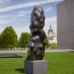 A large bronze sculpture that is oblong, stands upright, and appears lumpy sits on top of a concrete block in an outdoor space.