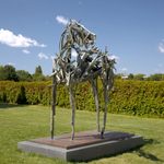A horse figure made of driftwood stands on a platform in a green garden space outside.