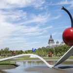 A giant red cherry sits on a spoon sculpture that overlooks a cathedral.