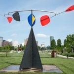 A large metal sculpture sits in a grassy lawn, a city view in the background. The sculpture has mobile-like arms with colorful objects on top.